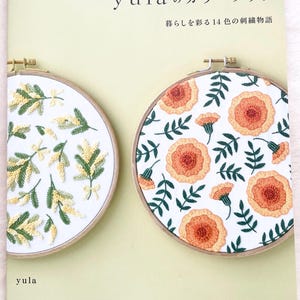 May include: A book titled "yula's Color Book: 14 Embroidery Stories to Color Your Life" with a green cover and two embroidery hoops. The hoops are on a light green background. The left hoop features a white fabric with yellow and green embroidery of mimosa flowers. The right hoop features a white fabric with orange and green embroidery of marigold flowers.