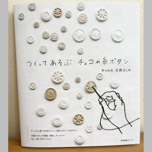 Puede incluir: Una portada de libro en japonés, titulada "Hacer y jugar con botones de hilo checos". La portada presenta varios botones de hilo beige y blanco sobre una tela blanca, con una mano bordada y texto en japonés.