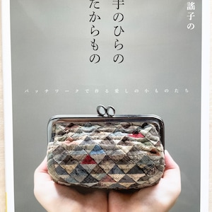 Può includere: Un libro intitolato "Mani di Tesori" con testo giapponese sulla copertina. Il libro tratta la creazione di piccoli oggetti in patchwork. La copertina presenta un primo piano di mani che tengono una piccola borsa trapuntata con una chiusura in argento.