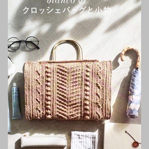 以下が含まれることがあります： ベージュとピンクの色合いでテクスチャーのあるヘリンボーン柄の長方形のクロッシェバッグ。2つの丸いハンドルが付いています。画像には日本語と英語のテキスト、丸いメガネ、折りたたみ傘、チューブ、ポーチ、布が含まれています。
