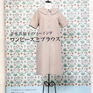 May include: A book cover featuring a beige dress with a white collar and black trim on a mannequin. The background is a floral pattern. The title of the book is "12 clothes by machiko kayaki" and the subtitle is "茅木真知子のソーイング ワンピースとブラウス".