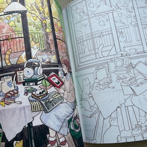 May include: A colouring book open to two pages showing a detailed illustration of a cosy indoor scene. One page is fully coloured with warm autumn tones, featuring a person reading at a table with cats, while the other page is a black and white line art.