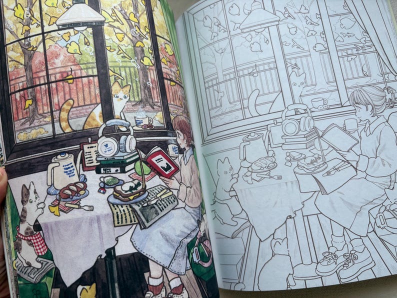 May include: A colouring book open to two pages showing a detailed illustration of a cosy indoor scene. One page is fully coloured with warm autumn tones, featuring a person reading at a table with cats, while the other page is a black and white line art.