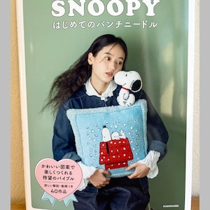 Op de afbeelding: Een boek getiteld "SNOOPY" in het Engels en Japans, met een vrouw die een blauw kussen vasthoudt met een Snoopy-ontwerp. Het kussen heeft een rood hondenhok en Snoopy-personages. De boekomslag is groen met Japanse tekst en een Snoopy-knuffel.