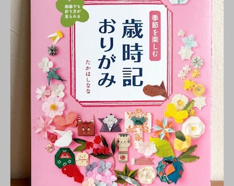 May include: A pink book cover titled "nanahoshi no 季節を楽しむ 歳時記おりがみ" featuring a variety of colorful origami figures arranged in a circular pattern. The book includes Japanese text and illustrations of folded paper flowers, animals, and seasonal items.