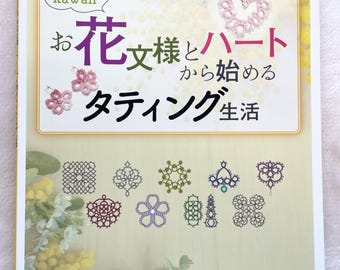 Puede incluir: Un libro en japonés titulado "Kawaii: O Hana Shi y Corazón, Tatting Life" con un diseño de tatting en forma de corazón rosa claro en la portada. La portada del libro también presenta un primer plano de una lanzadera de tatting y un fondo de flores de mimosa amarillas.