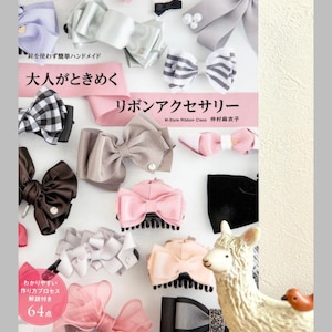 以下が含まれることがあります： ピンク、グレー、黒、白の色合いの手作りのリボンの例が掲載された「リボンアクセサリー」というタイトルの本。本の表紙には日本語のテキストが記載されています。装飾用のラマの置物も付いています。