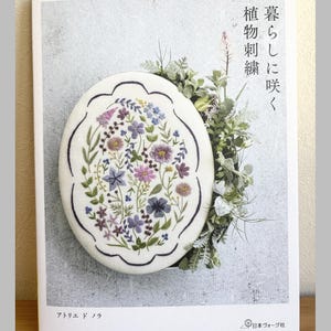 May include: A book cover with an embroidered floral design. The oval embroidery features purple, blue, and yellow flowers and green leaves. Japanese text is visible on the cover, along with the text "Pomadour's Craft Cafe".