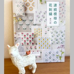 Peut inclure: Un livre japonais sur la broderie florale, avec le titre en japonais. Le livre présente divers motifs floraux brodés. Une figurine de mouton en céramique blanche avec un petit oiseau orange est devant le livre.