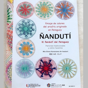 May include: A softcover instructional book titled ÑANDUTÍ featuring colourful circular lace patterns on the white cover. The text includes Spanish and Japanese characters, detailing Paraguayan lace techniques, with various intricate, multi-coloured circular designs displayed across the front.