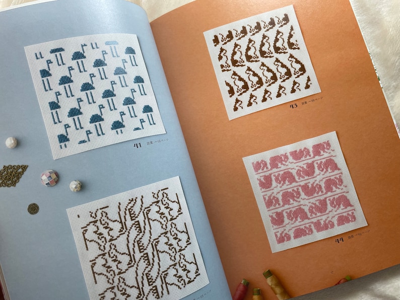 May include: A book with four different cross-stitch patterns. The patterns are on white fabric and feature different designs, including mushrooms, animals, and geometric shapes. The book has page numbers 41, 43, and 44.