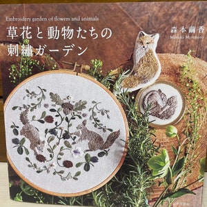 Puede incluir: Un libro titulado "Embroidery Garden of Flowers and Animals: 草花と動物たちの 刺繍ガーデン" de Mayuka Morimoto. La portada presenta un primer plano de un aro de bordado con un diseño de dos ardillas y bayas en una rama. El fondo muestra un zorro y una ardilla bordados en tela.