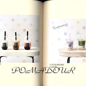 May include: Three miniature plastic cups with different colored drinks and tapioca pearls. The cups have black straws and are sitting on a white surface. The text "Yummy" is written in black above the cups. The text "タピオカ  珍珠" is written in black below the cups. The text "カラフルタピオカ  彩色珍珠" is written in black below the cups on the right side of the image.