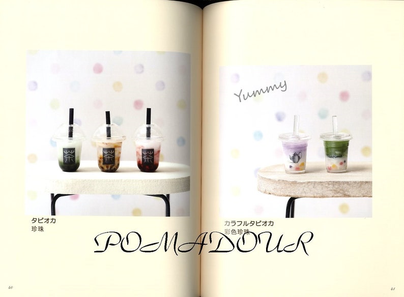 May include: Three miniature plastic cups with different colored drinks and tapioca pearls. The cups have black straws and are sitting on a white surface. The text "Yummy" is written in black above the cups. The text "タピオカ  珍珠" is written in black below the cups. The text "カラフルタピオカ  彩色珍珠" is written in black below the cups on the right side of the image.