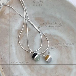 May include: Two silver chain necklaces with pendants. One pendant is a layered jade stone in shades of green, black, and white. The other pendant is a layered jade stone in shades of yellow, white, and gray. The necklaces are on a light gray background with the text "SERENE" and "HYPOALLERGENIC STERLING SILVER" and "ORIENTAL AESTHETIC JADE PENDANT" written on the background.