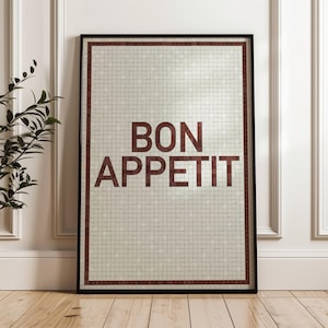 May include: Framed wall art with the words "BON APPETIT" in dark red letters. The artwork features a mosaic tile design in shades of white and beige, framed by a dark red border and a black frame. The art is displayed against a white wall.