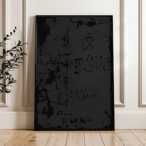 May include: A framed black artwork with the word "JESUS" and other illegible text scrawled across it. The artwork has a distressed, textured appearance, with a star symbol visible. The frame is black, and the artwork is displayed against a white wall.
