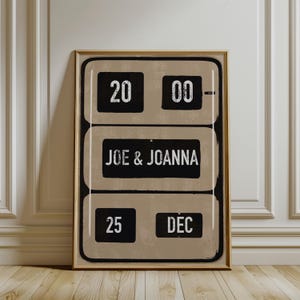 May include: Framed print featuring a vintage-style design. The print displays the numbers "20 00" at the top, the names "JOE & JOANNA" in the middle, and "25 DEC" at the bottom. The background is a neutral tan color, with black text and a gold frame.