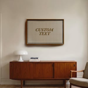 May include: A framed artwork with the words "CUSTOM TEXT" in gold lettering. The artwork is mounted on a white background and has a grid pattern. Below, a mid-century modern wooden cabinet with sliding doors and a white mushroom lamp.