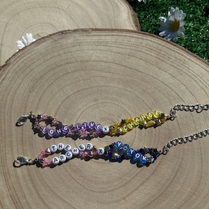 May include: Two beaded bracelets with silver clasps. The top bracelet is yellow, orange, and purple with the words "LONG LIVE X CHANGE". The bottom bracelet is pink, blue, and white with the words "THE ARCHER X YO YO K".