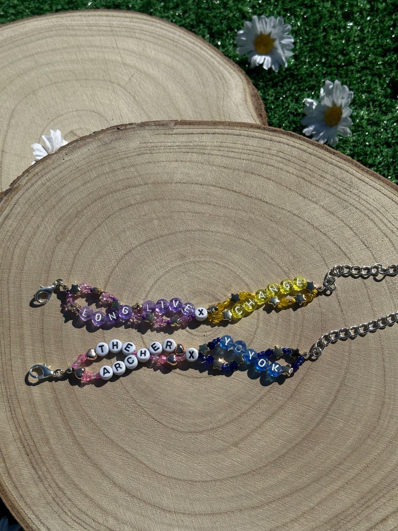 May include: Two beaded bracelets with silver clasps. The top bracelet is yellow, orange, and purple with the words "LONG LIVE X CHANGE". The bottom bracelet is pink, blue, and white with the words "THE ARCHER X YO YO K".