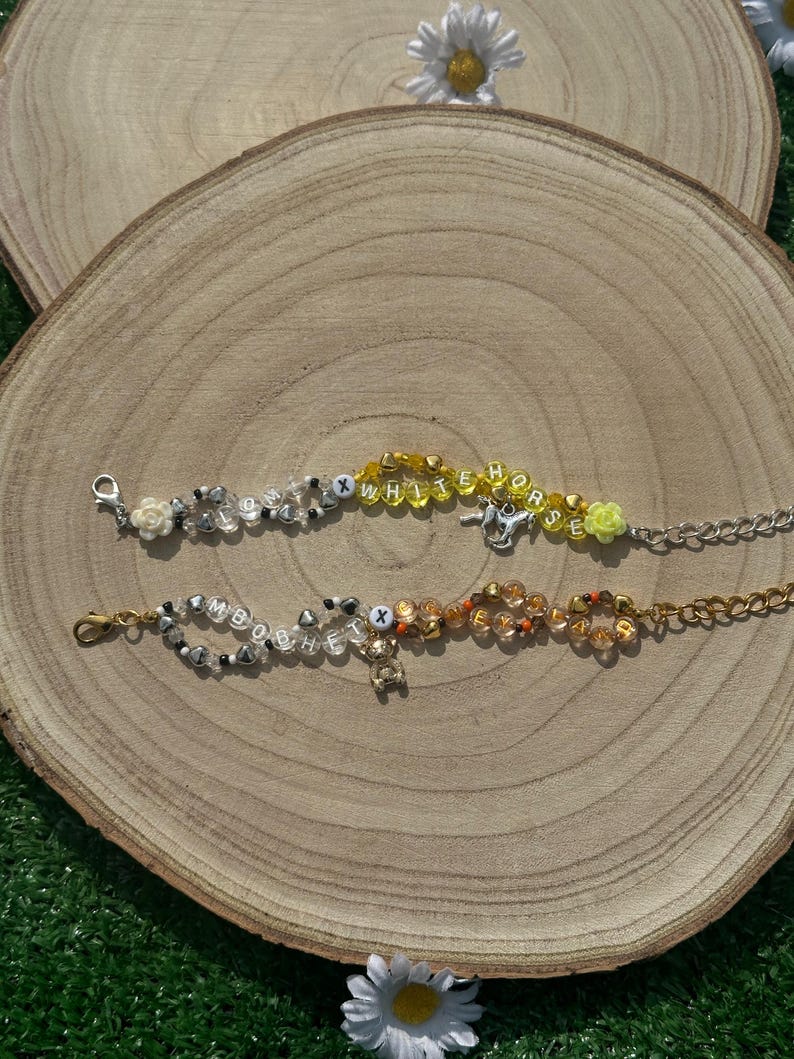 May include: Two beaded bracelets with charms. The top bracelet spells "WHITE HORSE" in yellow beads with silver accents. The bottom bracelet spells "BOBBY" in white beads with silver accents. Both bracelets have a silver clasp.