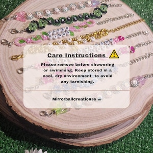 May include: A collection of colourful beaded bracelets with letter beads spelling out words like "Gorgeous", "Paper Rings", and "Love You". The bracelets are displayed on a wooden surface with a care instructions card that reads "Please remove before showering or swimming. Keep stored in a cool, dry environment to avoid any tarnishing. Mirrorballcreationss".