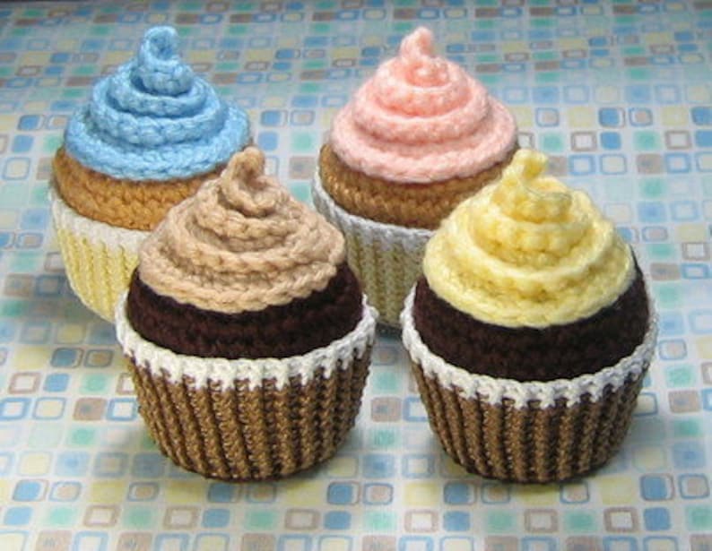 May include: Three crocheted cupcakes, one blue, one pink, and one yellow, with chocolate frosting and white sprinkles.
