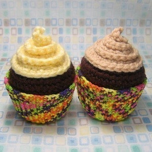 May include: Two crocheted cupcakes with chocolate frosting and colorful cupcake liners. One cupcake has a light yellow frosting swirl and the other has a light brown frosting swirl.