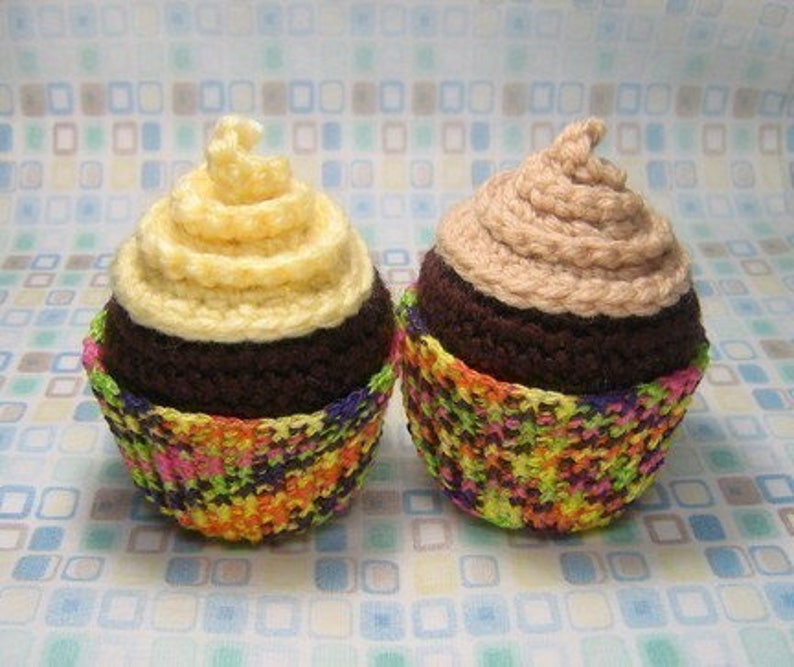 May include: Two crocheted cupcakes with chocolate frosting and colorful cupcake liners. One cupcake has a light yellow frosting swirl and the other has a light brown frosting swirl.