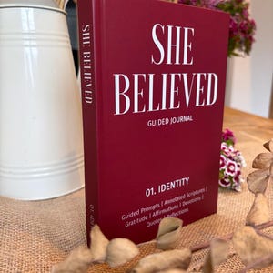 Może przedstawiać: Ciemnoczerwony dziennik w twardej oprawie z tytułem SHE BELIEVED napisanym białą czcionką szeryfową. Okładka zawiera podtytuł GUIDED JOURNAL, tekst 01. IDENTITY oraz listę funkcji, w tym Guided Prompts, Annotated Scriptures, Gratitude, Affirmations, Devotions, Quotes i Reflections.