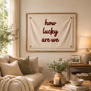 Può includere: Un arazzo rettangolare in tessuto color bianco sporco incorniciato in legno chiaro, con il testo ricamato in rosso scuro "how lucky are we" in corsivo. Il tessuto è fissato con quattro occhielli metallici color ottone agli angoli.