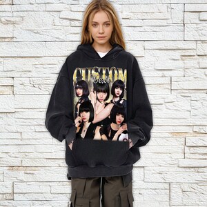 Pu&ograve; includere: Felpa con cappuccio nera con una grafica di un gruppo di donne con la scritta "Custom Text" in lettere dorate. La felpa ha un cappuccio con coulisse e una tasca frontale. La modella indossa la felpa.