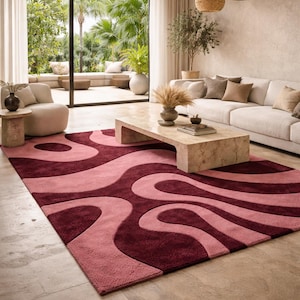 Puede incluir: Una alfombra burdeos y rosa con un patrón ondulado domina la habitación. La alfombra está colocada sobre un suelo de baldosas, y una mesa de centro de mármol se encuentra en el centro. La habitación tiene una paleta de colores neutros con vistas a un espacio exterior verde.