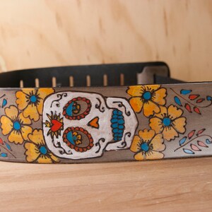 Puede incluir: Una correa de guitarra de cuero gris con un diseño de calavera de azúcar pintada. La calavera es blanca con contornos negros y presenta detalles azules, rojos y amarillos. La correa está decorada con flores amarillas y hojas verdes. El texto "Ars Nova" está escrito en la correa.