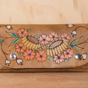 May include: A brown leather wallet with a floral design. The flowers are painted in shades of pink, orange, and yellow. The wallet has a flap closure and a silver snap.
