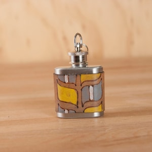 Puede incluir: Una petaca de plata con un patrón geométrico amarillo, marrón y gris en el exterior. La petaca tiene una tapa de plata y un pequeño lazo para sujetar una cadena.