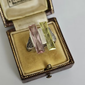 May include: A silver ring with a geometric design, featuring a pink and a yellow rectangular gemstone. The ring is displayed in a vintage brown jewelry box with a gold interior. The box has the text "By Appointment Vickery Bournemouth".