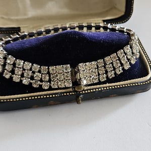 May include: A three-row rhinestone bracelet featuring clear, square-cut stones set in a silver-toned metal frame. The bracelet is displayed inside a rectangular, black-cased jewellery box with a dark blue velvet interior lining and a hinged lid.