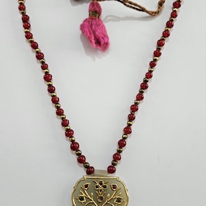 Puede incluir: Un collar con cuentas rojas y detalles dorados. El colgante tiene forma rectangular con un marco dorado y un diseño floral. Una borla rosa cuelga del cordón del collar.