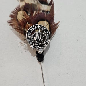 Puede incluir: Un pin de solapa plateado con un emblema circular con las palabras "ALTA PETE" y un diseño detallado. El pin está adornado con plumas marrones y beige, creando un accesorio decorativo.