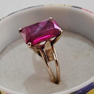 May include: A gold-toned ring featuring a large, rectangular, magenta-colored gemstone. The ring's band has a split design, and the gemstone is held in place by four prongs. The ring is set inside a floral patterned container.