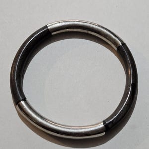 May include: A circular bracelet with a dark, possibly wooden, band and silver-colored metal accents. The bracelet is a complete circle, with the silver metal segments evenly spaced around the circumference. The bracelet is shown against a neutral background.