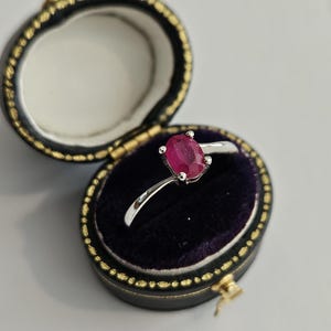 May include: A silver ring featuring a vivid red oval gemstone, held in place by four prongs. The ring is presented in an open, black, velvet-lined jewellery box with gold trim, set against a white backdrop.