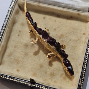 Op de afbeelding: Een goudkleurige hagedisbroche met donkerrode edelsteenaccenten, gepresenteerd in een vintage zwarte sieradendoos met een crèmekleurig interieur. Het lichaam van de hagedis is versierd met de edelstenen.