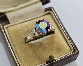 May include: A silver-toned ring featuring a round, faceted, iridescent stone that shifts between shades of deep purple and green. The ring band displays intricate, openwork metal detailing on the shoulders, presented in a dark brown box.