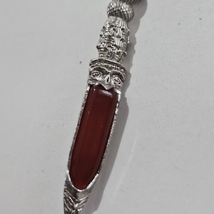 May include: A silver pendant with a rectangular, reddish-brown stone. The pendant has an ornate design featuring a face and other detailed elements. The pendant is approximately 7.6 cm long.