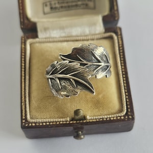 May include: A silver ring featuring two overlapping leaf designs with detailed veining and an oxidised finish, presented inside a brown rectangular jewellery box with a cream-coloured velvet interior and the text J. Vickery Bournemouth.