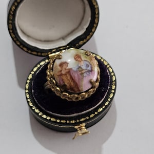 Peut inclure: Bague vintage avec une scène miniature peinte de personnages, dans un sertissage doré. La bague est présentée dans un écrin noir avec une bordure dorée et un intérieur en velours violet.