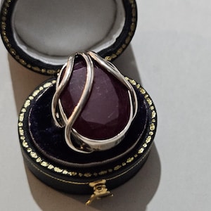 Può includere: Un grande pendente a goccia di pietra preziosa rosso scuro con un filo d'argento, presentato in un cofanetto nero con finiture dorate. Il pendente è incastonato su un interno in velluto blu scuro.